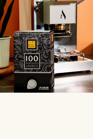 Boîte de 18 dosettes 100 Percento Arabica et machine à dosettes Aroma en arrière-plan.