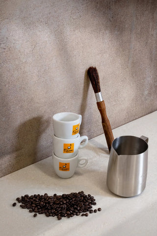 Tre tazzine da espresso Filicori Zecchini impilate. Lattiera professionale da cappuccino. Chicchi di caffè in primo piano.